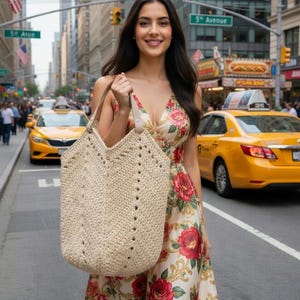 K&ouml;nnte beinhalten: Eine Frau in einem gebl&uuml;mten Kleid tr&auml;gt eine gro&szlig;e, beige geh&auml;kelte Tragetasche mit Ledergriffen. Das Kleid hat einen cremefarbenen Hintergrund mit roten Rosen und goldenen Akzenten. Die Szene spielt auf einer Stadtstra&szlig;e mit gelben Taxis.