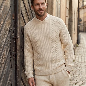 May include: A cream-colored cable knit sweater with a crew neckline and long sleeves, worn over a white collared shirt, paired with matching cream-colored knit trousers. The sweater features a textured pattern of vertical cables and honeycomb stitches.