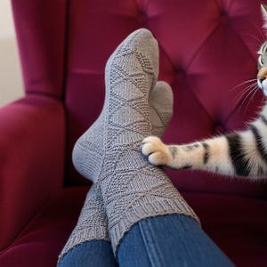 May include: A pair of gray knitted socks with a diamond pattern, worn with blue jeans. A cat's paw gently touches one sock. The socks are knee-high and appear soft and warm, perfect for cozy days.