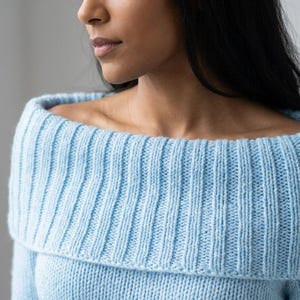 Può includere: Un maglione in maglia azzurro caratterizzato da un ampio colletto a coste ripiegato che poggia sulle spalle. Il capo è realizzato con un motivo a maglia testurizzato, che mostra un filato azzurro uniforme su tutto il pezzo.