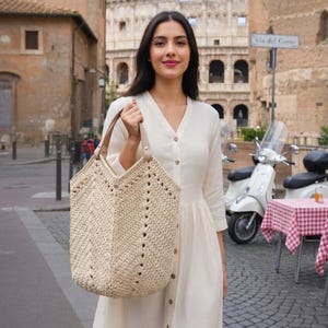 K&ouml;nnte beinhalten: Beige geh&auml;kelte Tragetasche mit braunen Ledergriffen. Die Tasche hat ein geometrisches Design mit kleinen L&ouml;chern. Die Frau tr&auml;gt ein wei&szlig;es Kleid. Der Hintergrund zeigt eine europ&auml;ische Stra&szlig;enszene.