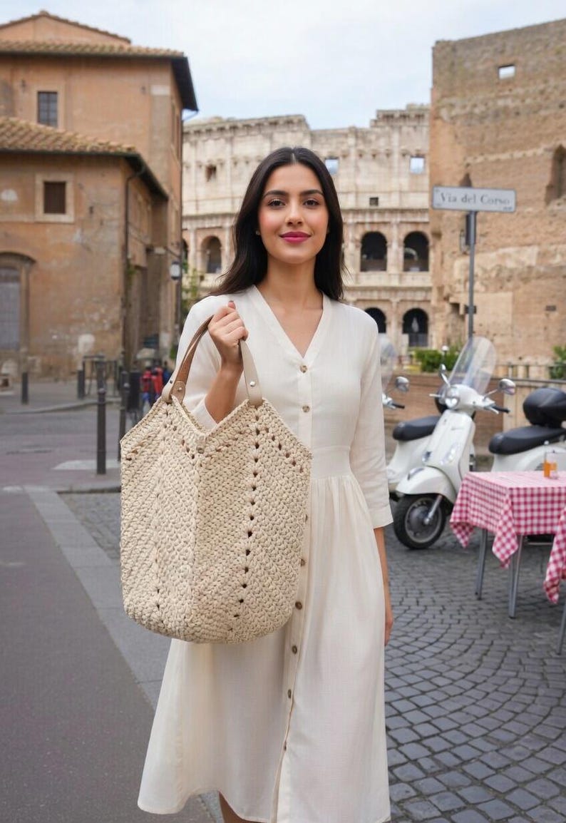 K&ouml;nnte beinhalten: Beige geh&auml;kelte Tragetasche mit braunen Ledergriffen. Die Tasche hat ein geometrisches Design mit kleinen L&ouml;chern. Die Frau tr&auml;gt ein wei&szlig;es Kleid. Der Hintergrund zeigt eine europ&auml;ische Stra&szlig;enszene.
