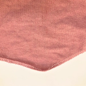 Pode incluir: Close-up de um tecido de malha rosa empoeirado com um padr&atilde;o de ponto vis&iacute;vel. Uma agulha de tric&ocirc; &eacute; inserida no tecido, com um pequeno la&ccedil;o de fio vis&iacute;vel. O tecido tem uma forma triangular, sugerindo um xale ou cachecol em andamento.