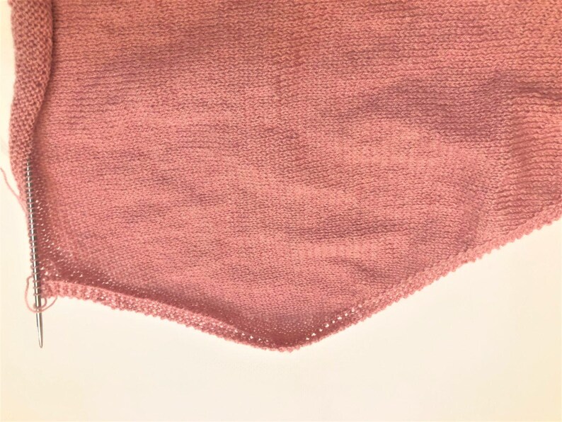 Pode incluir: Close-up de um tecido de malha rosa empoeirado com um padr&atilde;o de ponto vis&iacute;vel. Uma agulha de tric&ocirc; &eacute; inserida no tecido, com um pequeno la&ccedil;o de fio vis&iacute;vel. O tecido tem uma forma triangular, sugerindo um xale ou cachecol em andamento.