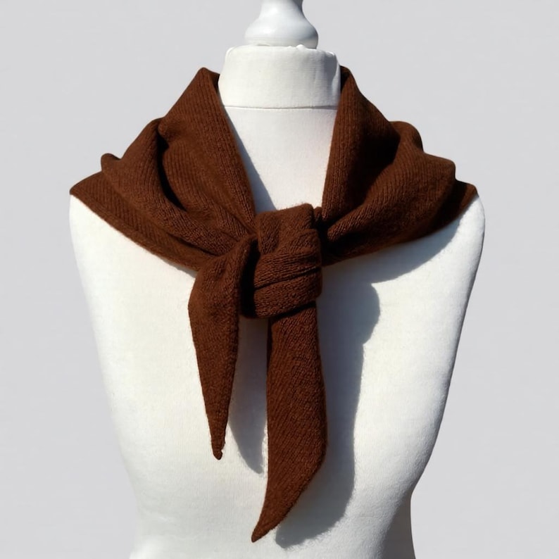 Pode incluir: Uma echarpe de malha castanha &eacute; drapeada sobre um manequim branco. A echarpe &eacute; atada na frente, com as pontas penduradas. A cor da echarpe &eacute; um castanho quente e rico, e a textura de malha &eacute; vis&iacute;vel. A echarpe parece ser feita de um material macio.