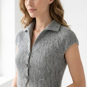 Könnte beinhalten: Eine graue Strickjacke mit kurzen Ärmeln, einem Kragen und einer Knopfleiste auf der Vorderseite mit drei sichtbaren Knöpfen. Das Kleidungsstück ist in einem strukturierten Rippenstrickmuster gefertigt und hat eine kurze Länge bis zur Taille.