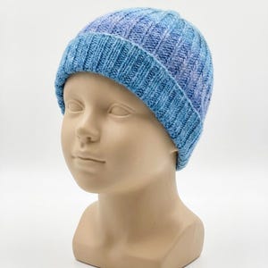 May include: A ribbed knit beanie in a variegated blue and light purple colorway. The hat features a thick, folded brim and a textured vertical stitch pattern throughout, crafted from a soft yarn blend that transitions between shades of blue and violet.
