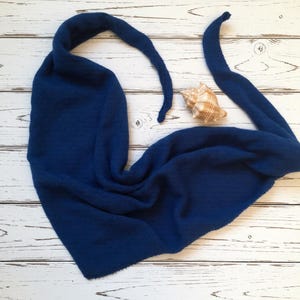 Pode incluir: Um cachecol azul marinho tricotado com pontas longas e pontiagudas, colocado sobre uma superf&iacute;cie de madeira branca. Uma concha marinha repousa perto do cachecol. A textura do cachecol &eacute; vis&iacute;vel, sugerindo um material macio e quente. A est&eacute;tica geral &eacute; simples e elegante.