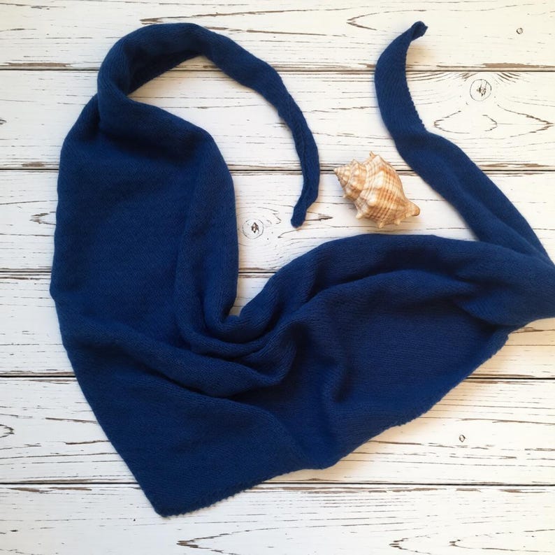 Pode incluir: Um cachecol azul marinho tricotado com pontas longas e pontiagudas, colocado sobre uma superf&iacute;cie de madeira branca. Uma concha marinha repousa perto do cachecol. A textura do cachecol &eacute; vis&iacute;vel, sugerindo um material macio e quente. A est&eacute;tica geral &eacute; simples e elegante.