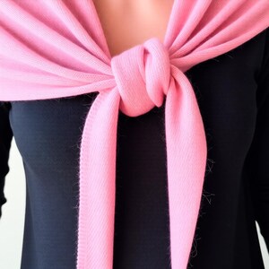 Pode incluir: Um cachecol rosa claro est&aacute; amarrado no pesco&ccedil;o, com as pontas penduradas. O cachecol est&aacute; sobre um top preto. O tecido macio do cachecol &eacute; vis&iacute;vel, com uma leve franja nas bordas.