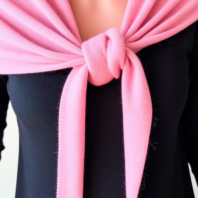 Pode incluir: Um cachecol rosa claro est&aacute; amarrado no pesco&ccedil;o, com as pontas penduradas. O cachecol est&aacute; sobre um top preto. O tecido macio do cachecol &eacute; vis&iacute;vel, com uma leve franja nas bordas.