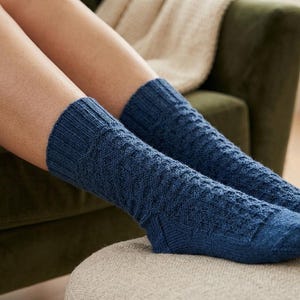 Puede incluir: Primer plano de calcetines de punto azul marino con un patrón texturizado. Los calcetines se llevan en las piernas de una persona, descansando sobre una superficie de color neutro. Los calcetines tienen un puño acanalado y un diseño detallado.