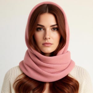 Pode incluir: Uma mulher usando um conjunto de capuz e cachecol de malha rosa claro. O capuz emoldura o rosto e o cachecol envolve o pesco&ccedil;o. A mulher est&aacute; vestindo um su&eacute;ter creme. O fundo &eacute; de uma cor neutra s&oacute;lida.