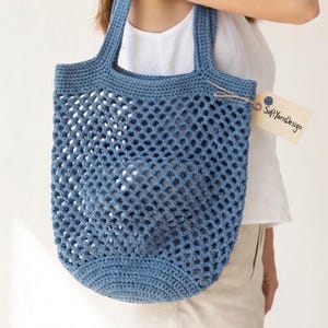 May include: A blue crocheted tote bag with a net-like design. The bag has two handles and a rounded bottom. A small tag with the text "Soft Yarn Design" is attached to the handle. The bag is made of a soft, flexible material.