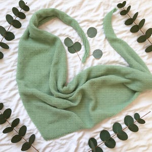 Pode incluir: Um cachecol de malha verde claro, drapeado sobre uma superf&iacute;cie branca, com um design longo e fluido. O cachecol &eacute; feito de um material macio e texturizado. Folhas de eucalipto dispostas ao redor do cachecol.