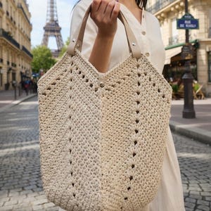 K&ouml;nnte beinhalten: Cremefarbene H&auml;keltasche mit braunen Ledergriffen. Die Tasche hat ein strukturiertes, offenes Webmuster und einen Knopfverschluss. Die Tasche wird vor dem Eiffelturm in Paris gehalten.