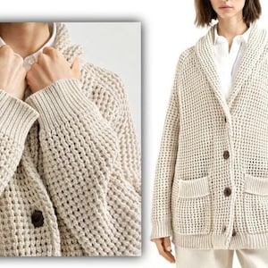 May include: Cream-colored, waffle-knit cardigan with a shawl collar and two front pockets. The sweater has a button-up closure with dark brown buttons. The cuffs and hem are ribbed. The cardigan is worn over a white collared shirt.