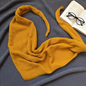 Pode incluir: Cachecol de malha amarelo mostarda sobre um cobertor cinza. O cachecol tem formato triangular com pontas longas e fluidas. Um livro e &oacute;culos com arma&ccedil;&atilde;o preta est&atilde;o por perto. A textura do cachecol sugere um material macio e quente.