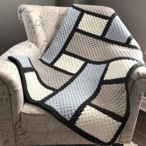 Può includere: Una coperta all'uncinetto fatta a mano con un motivo geometrico di quadrati e rettangoli nei toni del blu, bianco e grigio, bordata di nero. La coperta è drappeggiata su una sedia. Le cuciture strutturate suggeriscono un articolo morbido e fatto a mano.