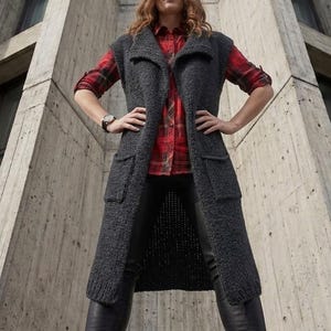 May include: A long, sleeveless charcoal gray knit vest featuring a wide collar, two large front patch pockets, and a textured wool-like finish. The vest is worn over a red and black plaid button-down shirt and black trousers with black boots.