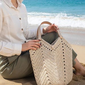 K&ouml;nnte beinhalten: Beige geh&auml;kelte Tragetasche mit V-f&ouml;rmigem Oberteil und Ledergriffen. Die Tasche hat ein strukturiertes, offen gewebtes Design und wird von einer Person gehalten, die an einem Sandstrand sitzt. Der Hintergrund zeigt das Meer und den blauen Himmel.