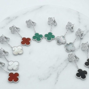 Peut inclure: Boucles d'oreilles pendantes argentées avec un motif de trèfle à quatre feuilles. Chaque boucle d'oreille est ornée d'un dessus serti de pierres précieuses et d'un pendentif en forme de trèfle de différentes couleurs : rouge, vert, blanc et noir. Présentées sur une surface en marbre blanc.