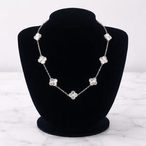 Op de afbeelding: Een zilveren ketting met zeven witte klavervormige hangers. De ketting wordt getoond op een zwarte mannequin tegen een witte en grijze marmeren achtergrond. De klaverhangers zijn gelijkmatig verdeeld over de ketting.