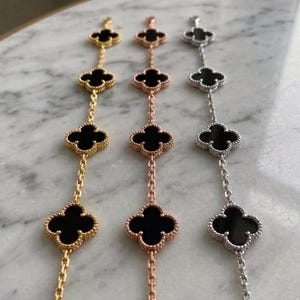 Op de afbeelding: Drie armbanden met klavervormige bedels. Eén is goudkleurig, één is roségoudkleurig en de derde is zilverkleurig. Elke bedel heeft een zwart midden en is verbonden door een fijne ketting.
