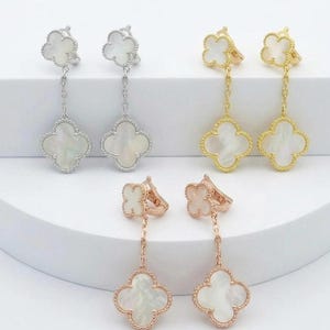 May include: A selection of four-leaf clover drop earrings in silver, gold, and rose gold. Each earring has a clover-shaped stud and a dangling clover pendant with a white, pearlescent centre. The earrings are presented on a white surface.
