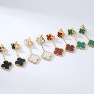 Puede incluir: Una colección de pendientes colgantes dorados, cada uno con un diseño de trébol de cuatro hojas. Los pendientes muestran varios colores, incluyendo negro, blanco, rojo y verde. Los colgantes de trébol cuelgan de tachuelas doradas.