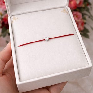 Può includere: Un braccialetto con filo rosso e un piccolo ciondolo a forma di cuore argentato, presentato in un portagioie bianco aperto. La scatola è tenuta in mano, con uno sfondo floreale sfocato. Il braccialetto è un design semplice ed elegante.