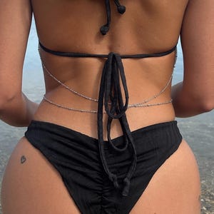 May include: A black bikini set featuring a halter neck top and ruched bottoms. A silver chain belt is worn around the waist. The person is seen from behind, with the sea in the background.