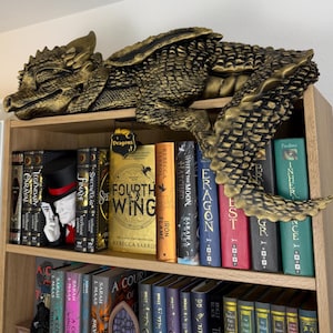 May include: A gold-coloured dragon ornament sits on a wooden bookshelf, above a collection of books. The dragon has detailed scales and wings. The books have colourful spines, with titles such as "Fourth Wing" and "Eragon".