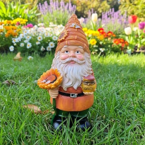 Puede incluir: Una estatua de gnomo de jardín con una larga barba blanca, sosteniendo un girasol y un tarro de miel. El gnomo viste un atuendo naranja, un sombrero marrón con un girasol y una abeja, y botas negras. El fondo presenta hierba verde y flores coloridas.