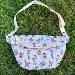 May include: White fanny pack with a gold zipper and cream-colored adjustable strap. The bag is patterned with colorful cartoon characters. The fanny pack is laying on a bed of green grass.