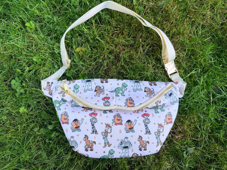 May include: White fanny pack with a gold zipper and cream-colored adjustable strap. The bag is patterned with colorful cartoon characters. The fanny pack is laying on a bed of green grass.