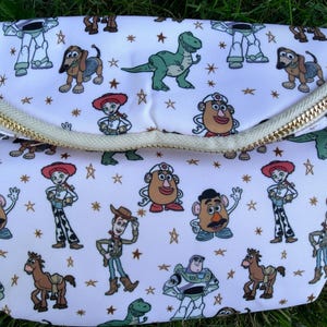 May include: White fanny pack with a gold zipper and a repeating pattern of cartoon characters. The characters include a green dinosaur, a brown dog, and a cowboy. The bag is on a green surface.
