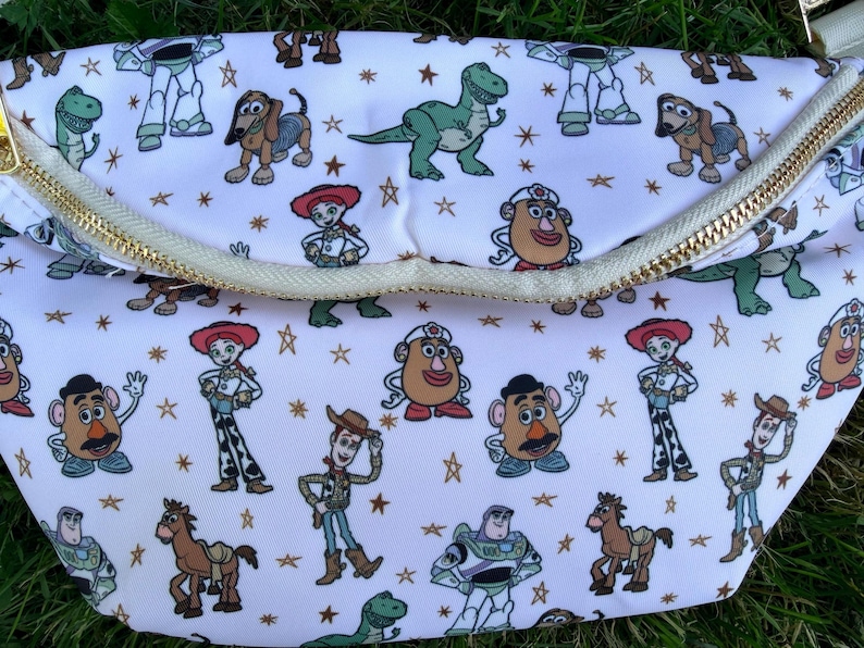 May include: White fanny pack with a gold zipper and a repeating pattern of cartoon characters. The characters include a green dinosaur, a brown dog, and a cowboy. The bag is on a green surface.