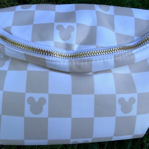 May include: A white and tan checkered fanny pack with a gold zipper. The bag features Mickey Mouse silhouettes in the tan squares. The zipper runs across the top of the bag.