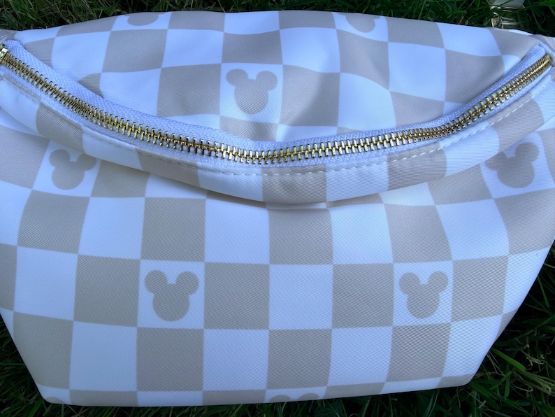 May include: A white and tan checkered fanny pack with a gold zipper. The bag features Mickey Mouse silhouettes in the tan squares. The zipper runs across the top of the bag.