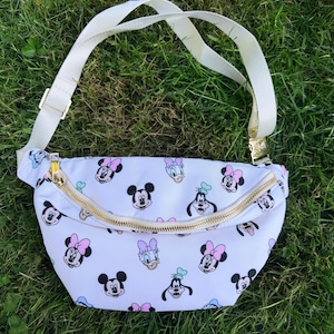 May include: White fanny pack with a gold zipper and adjustable white strap. The bag is patterned with images of Mickey Mouse, Minnie Mouse, Donald Duck, and Goofy. The bag is sitting on green grass.