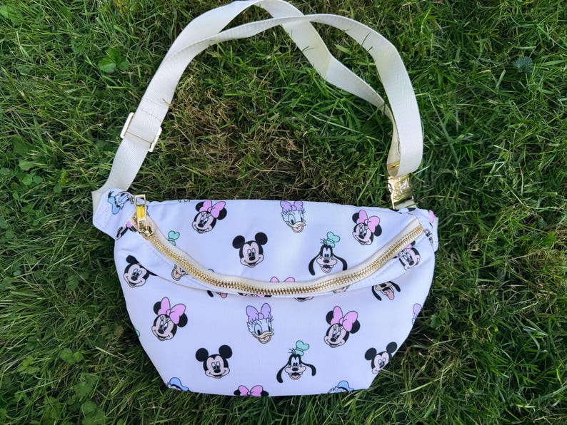 May include: White fanny pack with a gold zipper and adjustable white strap. The bag is patterned with images of Mickey Mouse, Minnie Mouse, Donald Duck, and Goofy. The bag is sitting on green grass.