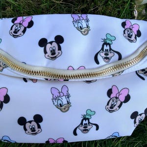 May include: White fanny pack with a gold zipper and a repeating pattern of Mickey Mouse, Minnie Mouse, Donald Duck, and Goofy. The characters are in black, pink, and blue on the white background.