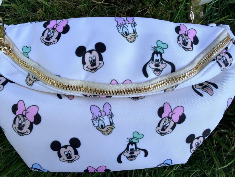 May include: White fanny pack with a gold zipper and a repeating pattern of Mickey Mouse, Minnie Mouse, Donald Duck, and Goofy. The characters are in black, pink, and blue on the white background.