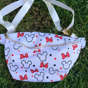 May include: White fanny pack with a gold zipper, featuring a pattern of black Mickey Mouse silhouettes, red bows, and red stars. The adjustable strap is white.