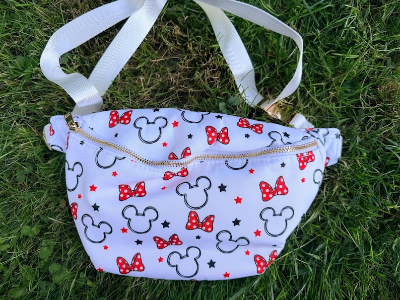 May include: White fanny pack with a gold zipper, featuring a pattern of black Mickey Mouse silhouettes, red bows, and red stars. The adjustable strap is white.