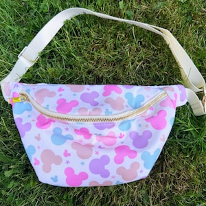 May include: A white fanny pack with a repeating pattern of pink, purple, and blue Mickey Mouse silhouettes and stars. The bag has a gold zipper and a cream-colored adjustable strap.