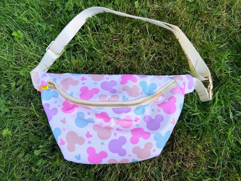 May include: A white fanny pack with a repeating pattern of pink, purple, and blue Mickey Mouse silhouettes and stars. The bag has a gold zipper and a cream-colored adjustable strap.