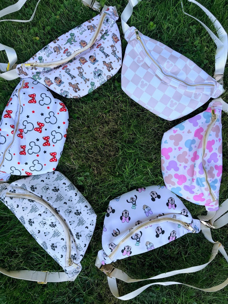 May include: A collection of colorful fanny packs with various patterns, including cartoon characters, Mickey Mouse shapes, and a pink and white checkered design. Each pack has a gold zipper and a white adjustable strap, set against a backdrop of green grass.