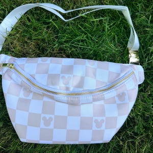 May include: A white and tan checkered fanny pack with a gold zipper and strap hardware. The bag features small Mickey Mouse silhouettes on the checkered pattern. The adjustable strap is white.